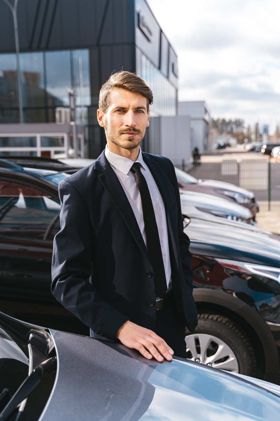 Professional man in a suit standing next to cars outside a modern building.